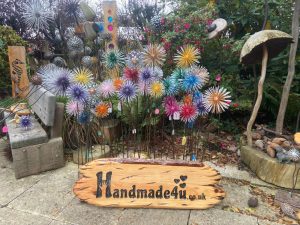 Metal garden ornament shaped like a dandelion flower, with delicate, radiating spokes resembling seeds blowing in the wind, standing in a garden bed surrounded by greenery